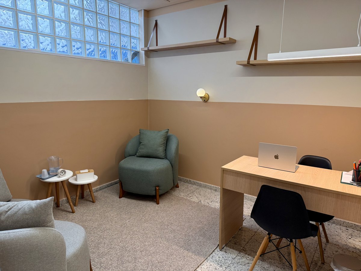 Consultório com poltrona verde, sofá, mesa de trabalho e luz natural pela janela de tijolos de vidro